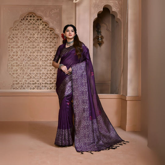 Woman wearing a purple silk saree with intricate geometric pallu and border, paired with traditional jewelry, standing in a heritage-inspired room with ornate carved walls and decorative window lattice.