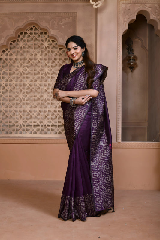 Woman wearing a purple silk saree with intricate geometric pallu and border, paired with traditional jewelry, standing in a heritage-inspired room with ornate carved walls and decorative window lattice.