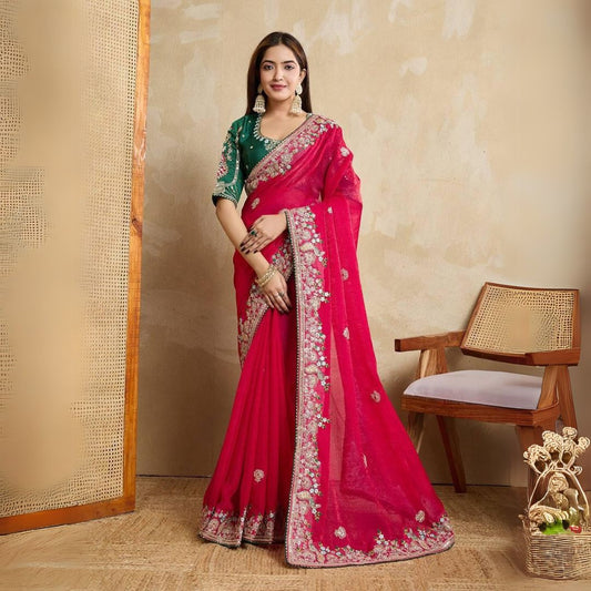 Woman wearing a royal purple silk saree with intricate pink and gold border and pallu, paired with a matching blouse and traditional gold jewelry, standing on a terracotta-tiled outdoor terrace with decorative plants and peach-colored walls