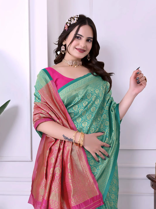 Woman wearing a teal silk saree with intricate gold motifs and a pink blouse, accessorized with traditional jewelry, posing indoors beside green plants and a wooden console table with decor items