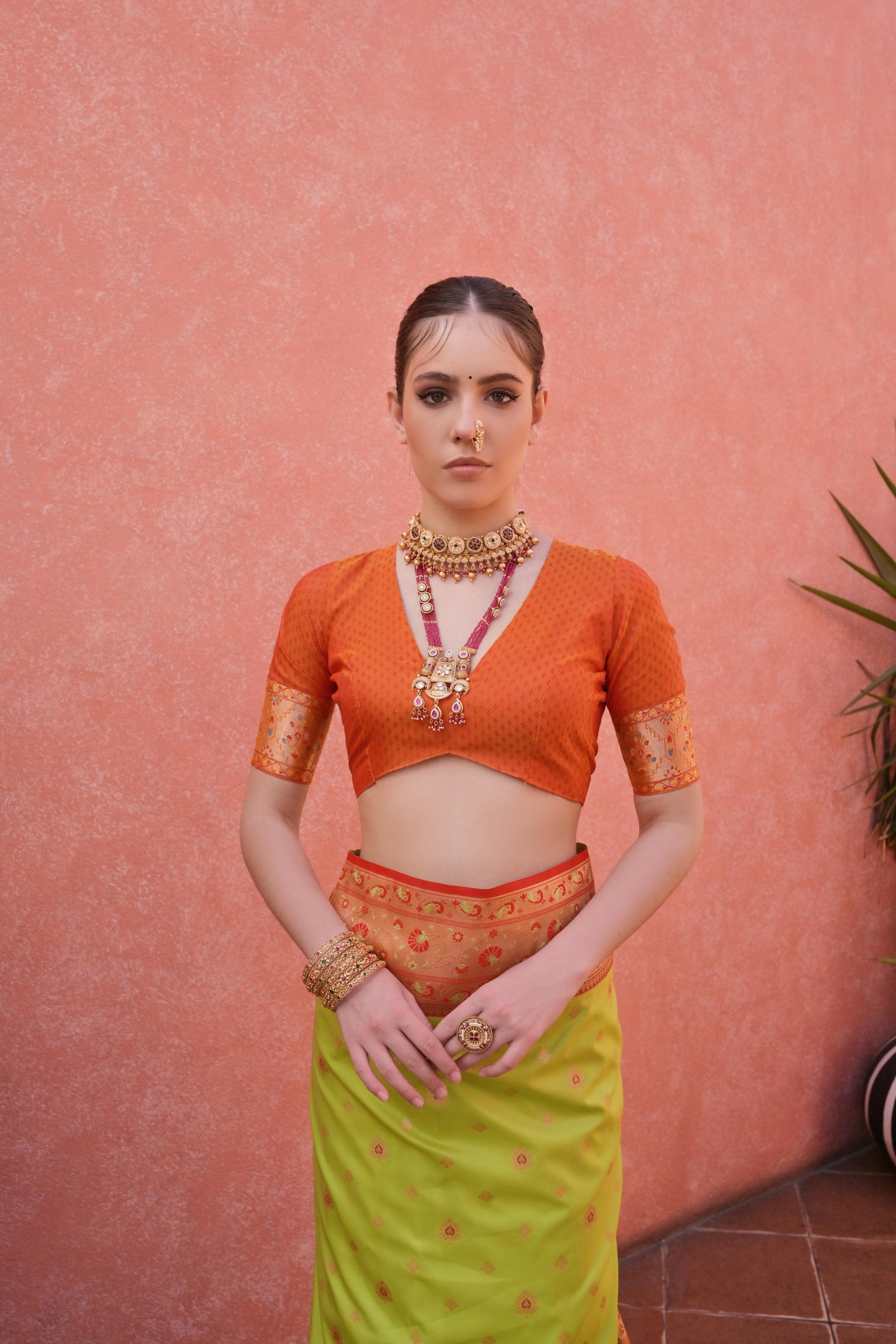 Woman wearing a green and orange traditional saree with matching blouse and jewelry, standing in a stylish courtyard with potted plants and pink walls