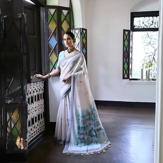 Woman wearing a white silk saree with pastel village motifs and a tassel border, paired with a light blue blouse and statement necklace, standing indoors beside vintage stained glass windows.