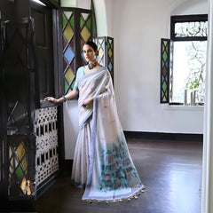 A woman models a graceful Off White Soft Muga Cotton Silk Saree. The pallu features a wide sea green panel decorated with traditional Warli-style figurative motifs in pink and red. She wears a light blue blouse and a dark oxidized choker necklace, posing indoors.