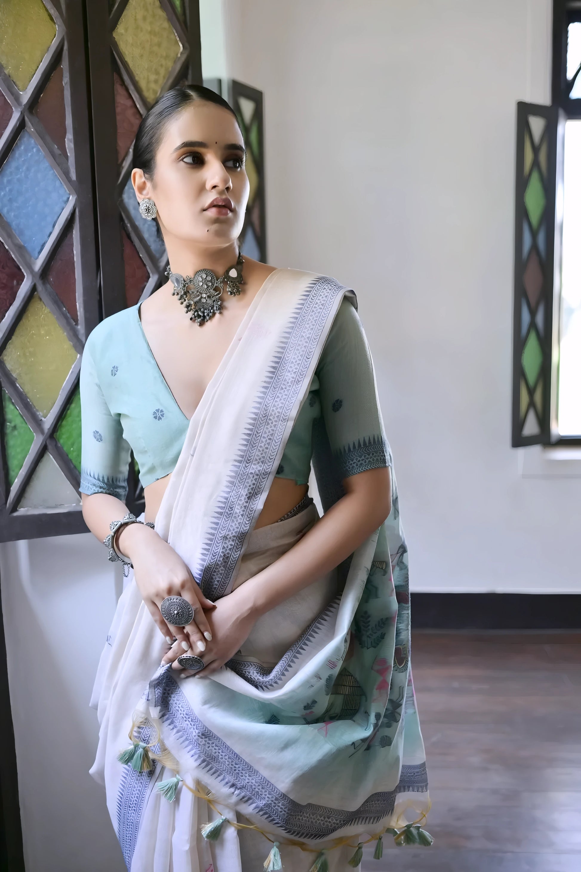 Woman wearing a white silk saree with pastel village motifs and a tassel border, paired with a light blue blouse and statement necklace, standing indoors beside vintage stained glass windows.