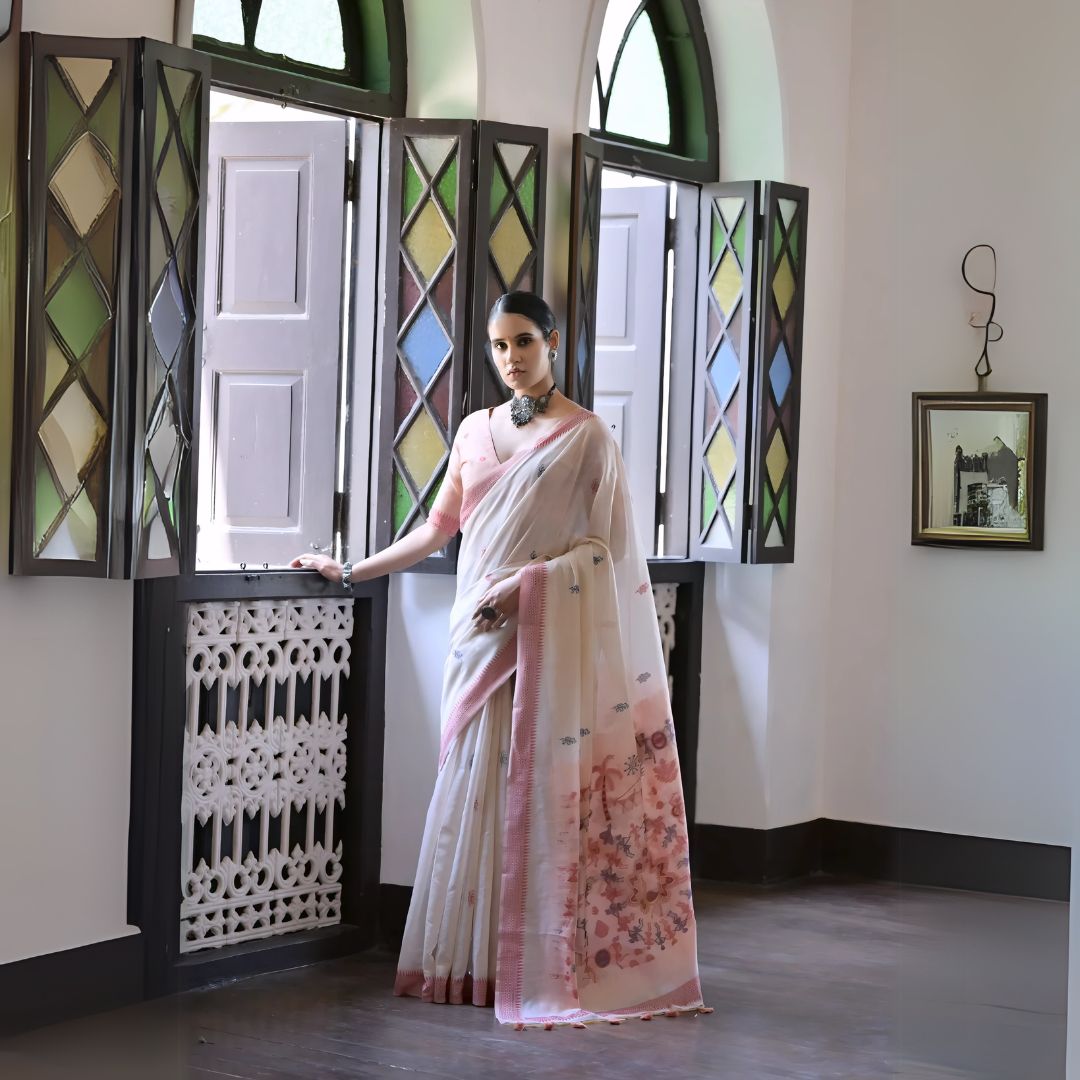 A woman models a graceful Off White Soft Muga Cotton Silk Saree. The saree has a dark pink woven border, and the pallu features a wide sky blue panel decorated with traditional Warli-style figurative motifs. She wears a light blue blouse and a dark oxidized choker necklace, posing indoors.