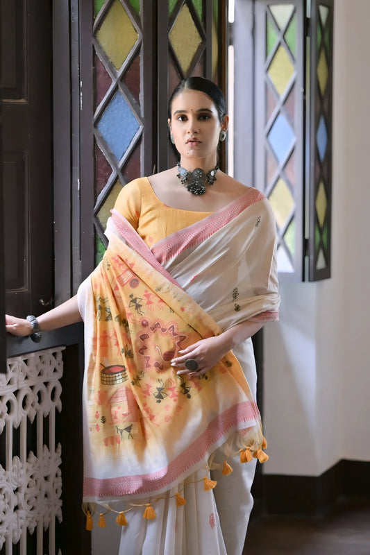 Woman wearing a white silk saree with pastel village motifs and a tassel border, paired with a yellow blouse and statement jewelry, standing indoors beside vintage stained glass windows