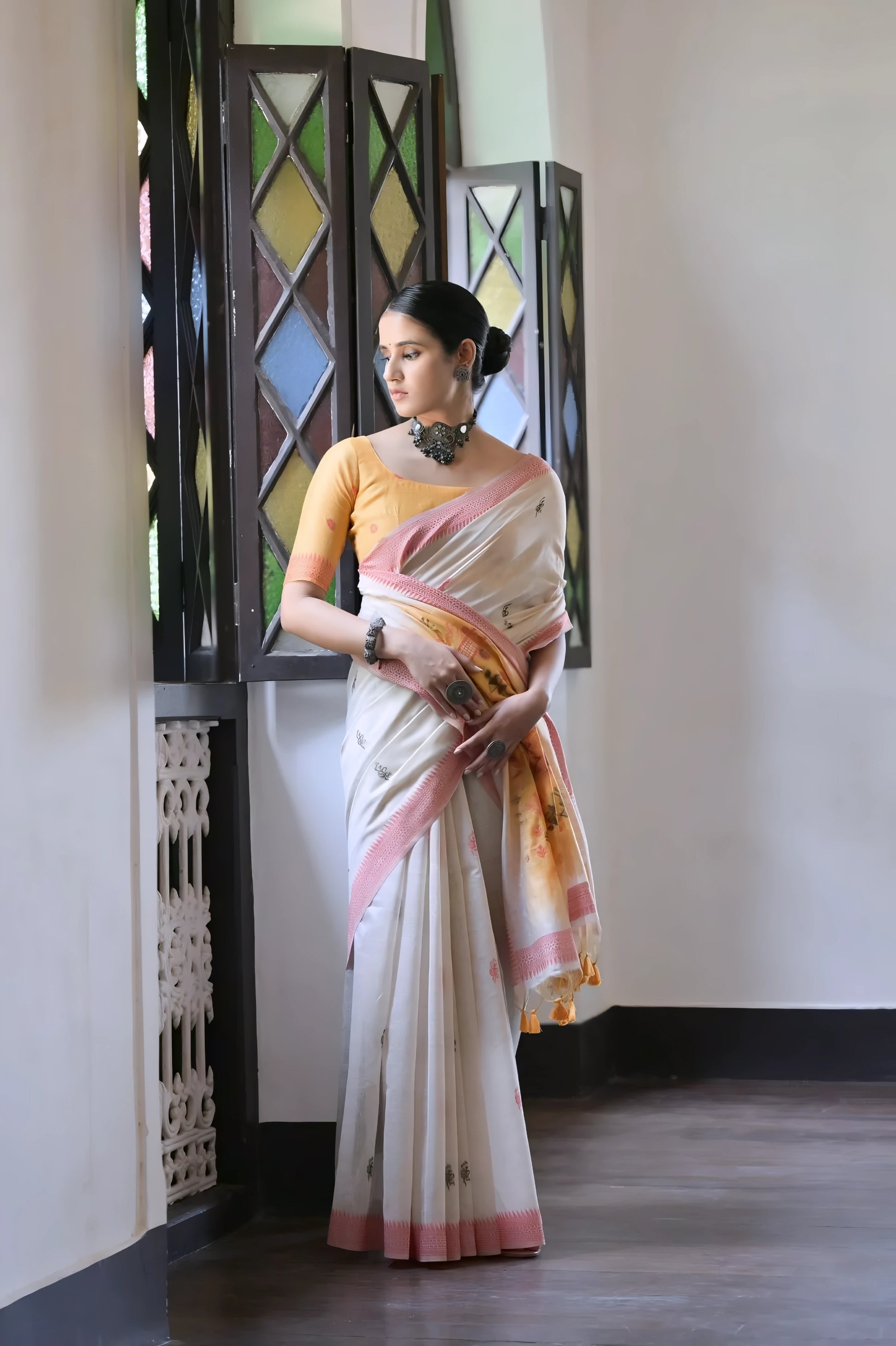 Woman wearing a white silk saree with pastel village motifs and a tassel border, paired with a yellow blouse and statement jewelry, standing indoors beside vintage stained glass windows.