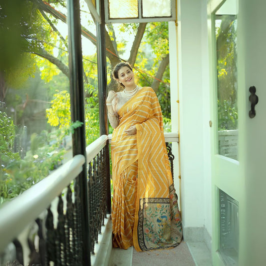 A woman models a vibrant Golden Yellow Saree with a white chevron/striped pattern. The pallu is beige with a dark border, featuring elaborate printed motifs of birds and flowers. She wears a pearl necklace and a lace-sleeve blouse, posing on a sunny balcony.