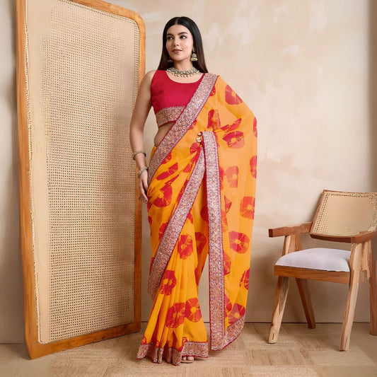 Woman wearing a yellow georgette saree with red floral patterns and an embellished pink border, paired with a sleeveless pink blouse and traditional jewelry, standing beside a wooden cane screen and modern chair against a beige wall.