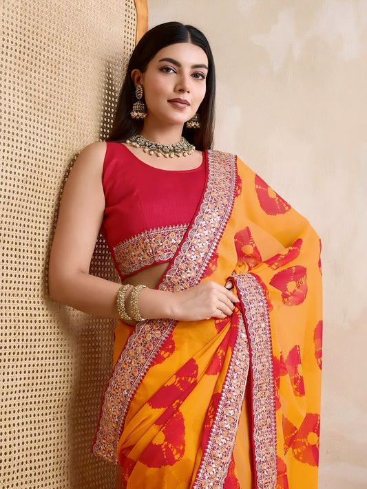 Woman wearing a yellow georgette saree with red floral patterns and an embellished pink border, paired with a sleeveless pink blouse and traditional jewelry, standing beside a wooden cane screen and modern chair against a beige wall.