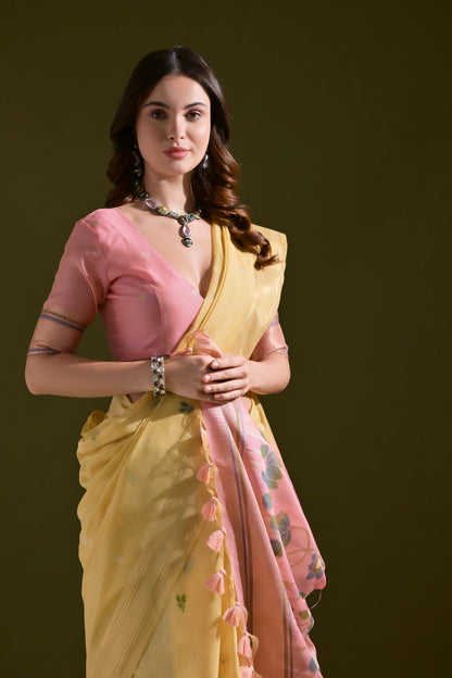 Woman wearing a yellow silk saree with pink floral pallu and tassel border, paired with a pink blouse and silver jewelry, standing against an olive green background.