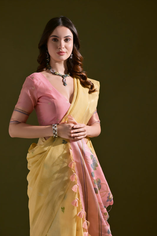 Woman wearing a yellow silk saree with pink floral pallu and tassel border, paired with a pink blouse and silver jewelry, standing against an olive green background.
