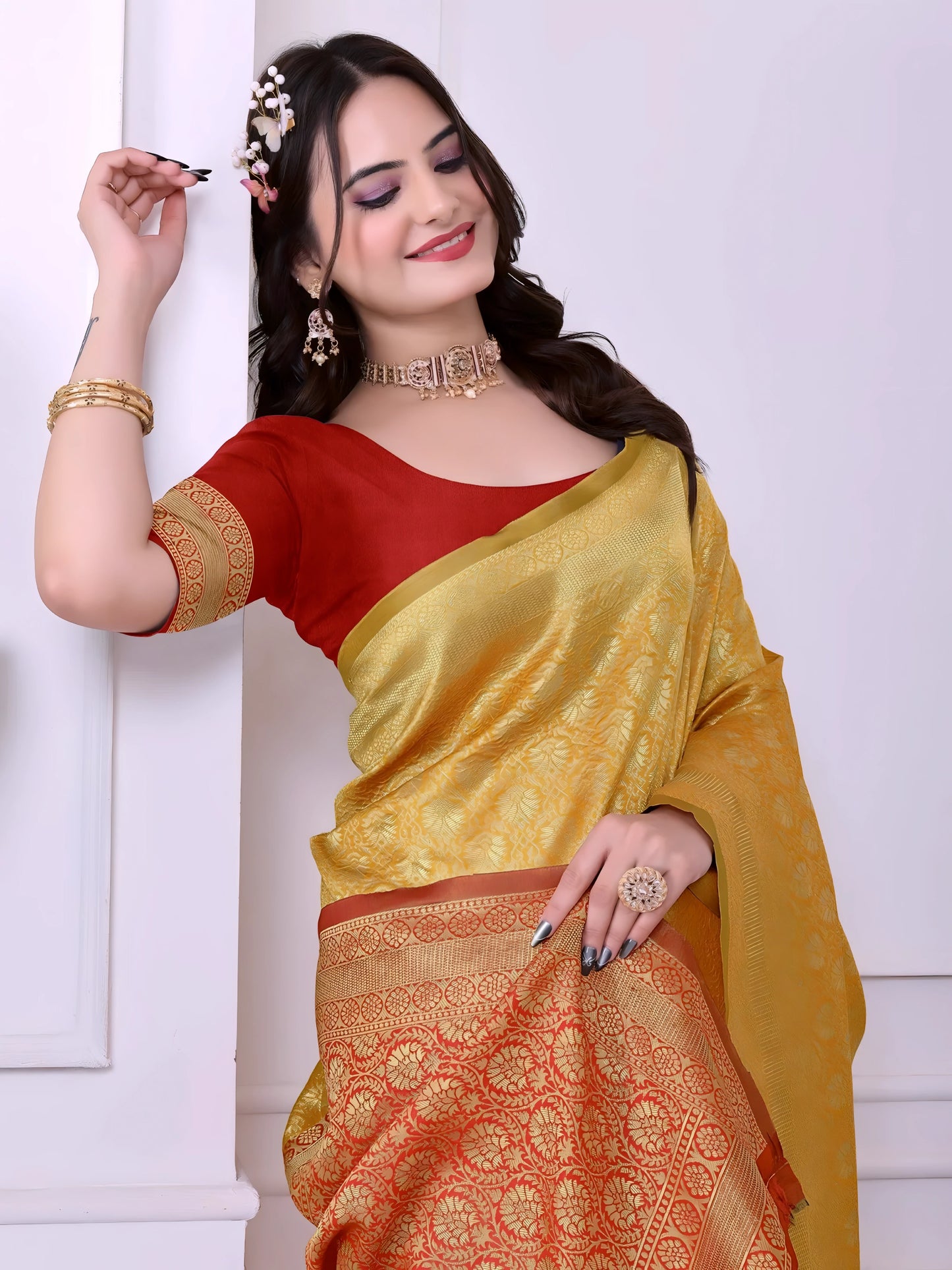  Woman wearing a yellow silk saree with intricate golden motifs and a contrasting red border, paired with a red blouse and traditional jewelry, standing indoors beside a potted plant and wooden console table.