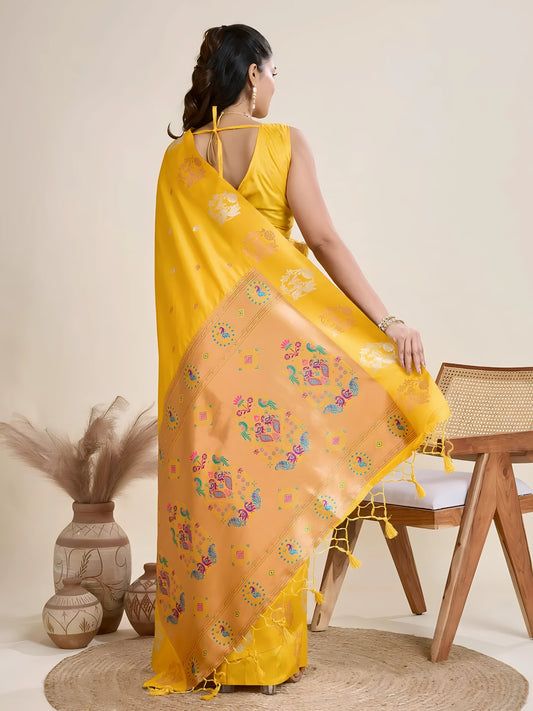 Woman wearing a vibrant yellow Banarasi silk saree with intricate pastel floral motifs and a contrasting gold border, accessorized with a statement necklace and bangles, elegantly posing beside a wooden chair in a minimalist studio setting with neutral decor, ceramic vases, and pampas grass.