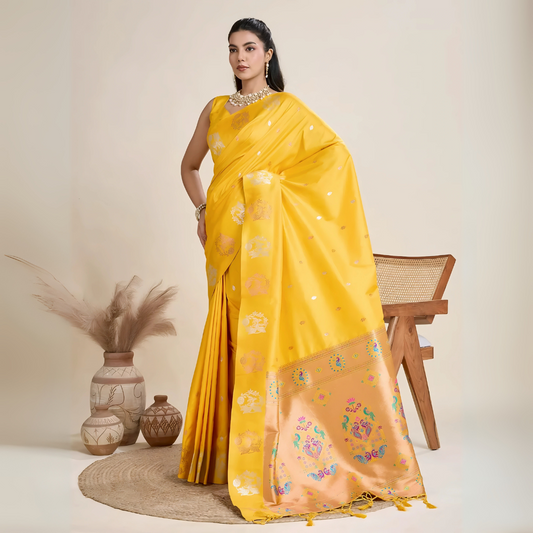 Woman wearing a vibrant yellow Banarasi silk saree with intricate pastel floral motifs and a contrasting gold border, accessorized with a statement necklace and bangles, elegantly posing beside a wooden chair in a minimalist studio setting with neutral decor, ceramic vases, and pampas grass.