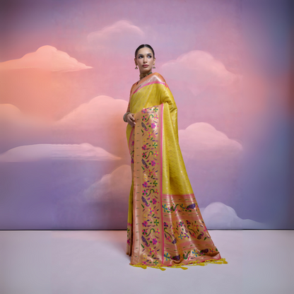 Woman wearing a yellow Banarasi silk saree with intricate pink and multicolor floral zari border and pallu, paired with traditional jewelry, posing in a pastel-themed studio with a dreamy cloud background