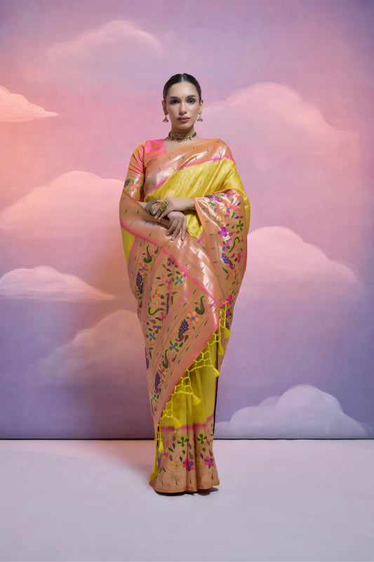 Woman wearing a yellow Banarasi silk saree with intricate pink and multicolor floral zari border and pallu, paired with traditional jewelry, posing in a pastel-themed studio with a dreamy cloud background