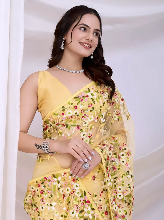 Woman wearing a yellow net saree with intricate multicolor floral embroidery and a matching sleeveless blouse, posing in a modern white studio with draped curtains and floral arrangements.
