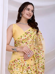 Woman wearing a yellow net saree with intricate multicolor floral embroidery and a matching sleeveless blouse, posing in a modern white studio with draped curtains and floral arrangements.