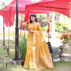 Woman wearing a yellow organza saree with delicate floral embroidery and a matching blouse, posing outdoors near a black lamp post under red canopies, with garden benches and vibrant greenery in the background.