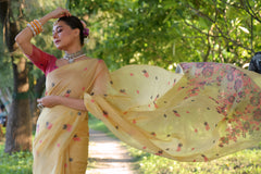 Woman wearing a yellow organza saree with delicate floral embroidery and a pink blouse, accessorized with traditional jewelry and bangles, standing on a sunlit garden path surrounded by lush green trees and foliage.