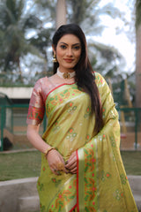 Woman wearing a yellow-green Banarasi silk saree with gold zari motifs and a vibrant pink contrast pallu featuring intricate temple and floral patterns, paired with a matching short sleeve blouse and traditional gold jewelry. She stands elegantly on a grassy lawn bordered by garden landscaping, with a background of fencing, lush trees, and playful outdoor structures, creating a graceful outdoor setting.