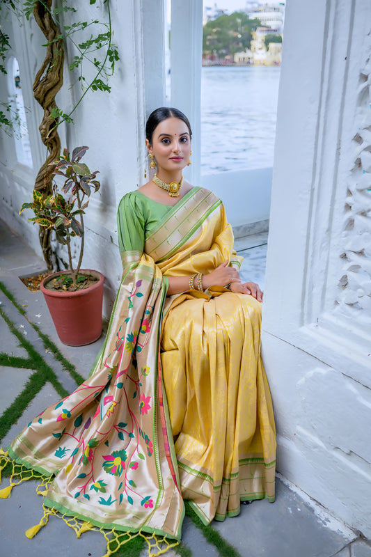 Woman wearing a yellow silk saree with green blouse and gold border, featuring a handpainted pallu with peacock and floral design, posing in a white heritage courtyard with potted plants and traditional architectural details