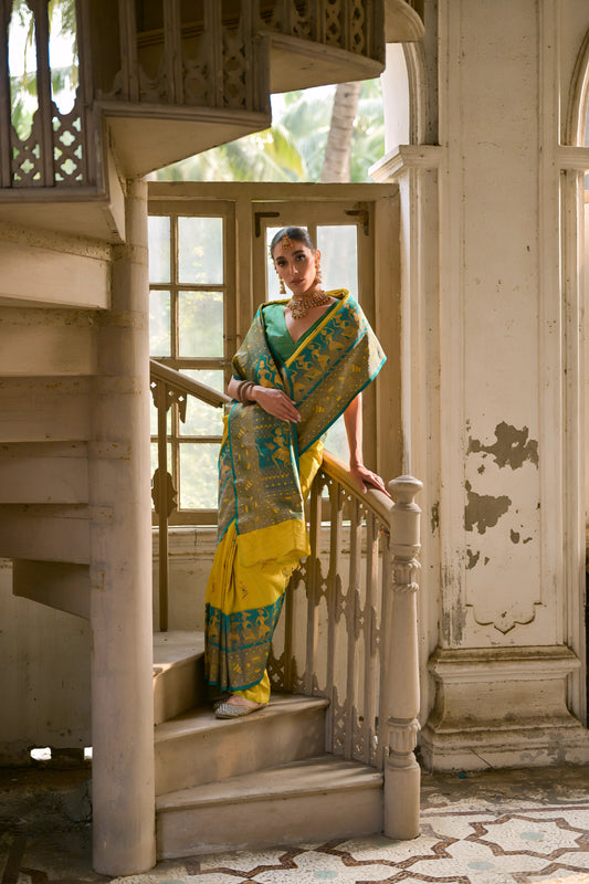 Woman wearing a bright yellow and green silk saree with intricate jacquard patterns and a green blouse, accessorized with traditional jewelry, gracefully standing on a vintage spiral staircase in a sunlit, historic interior featuring large wooden windows, ornate railings, distressed white walls, and geometric tiled flooring.