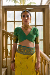 Woman wearing a bright yellow and green silk saree with intricate jacquard patterns and a green blouse, accessorized with traditional jewelry, gracefully standing on a vintage spiral staircase in a sunlit, historic interior featuring large wooden windows, ornate railings, distressed white walls, and geometric tiled flooring.