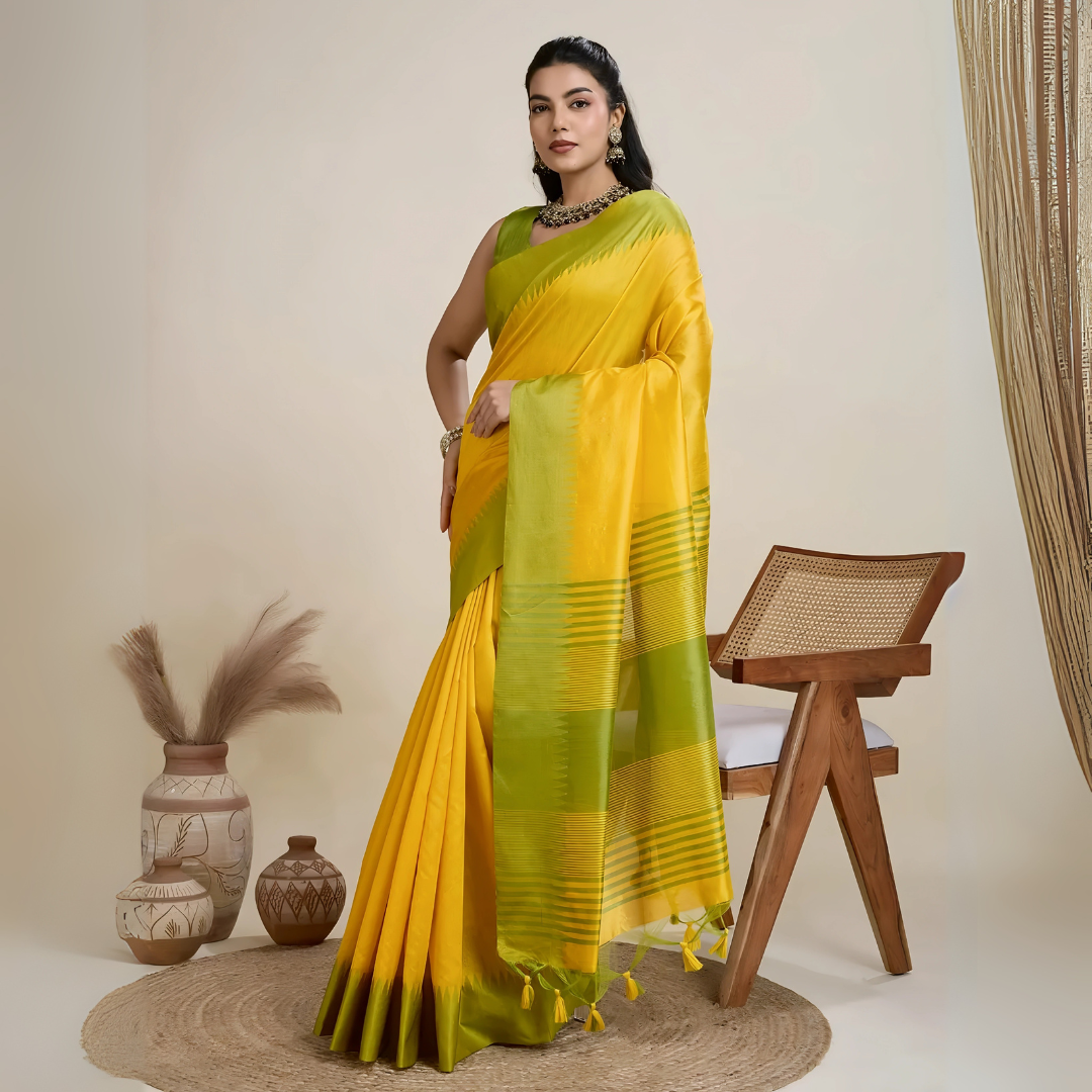 Woman wearing a yellow silk saree with a contrasting green border and pallu, paired with a matching blouse and traditional jewelry, elegantly posing beside a wooden chair in a minimalist studio setting with neutral decor, ceramic vases, and pampas grass.