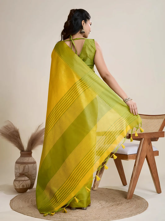 Woman wearing a yellow silk saree with a contrasting green border and pallu, paired with a matching blouse and traditional jewelry, elegantly posing beside a wooden chair in a minimalist studio setting with neutral decor, ceramic vases, and pampas grass.