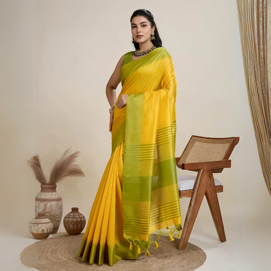 Woman wearing a yellow silk saree with a contrasting green border and pallu, paired with a matching blouse and traditional jewelry, elegantly posing beside a wooden chair in a minimalist studio setting with neutral decor, ceramic vases, and pampas grass.