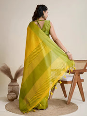 Woman wearing a yellow silk saree with a contrasting green border and pallu, paired with a matching blouse and traditional jewelry, elegantly posing beside a wooden chair in a minimalist studio setting with neutral decor, ceramic vases, and pampas grass.