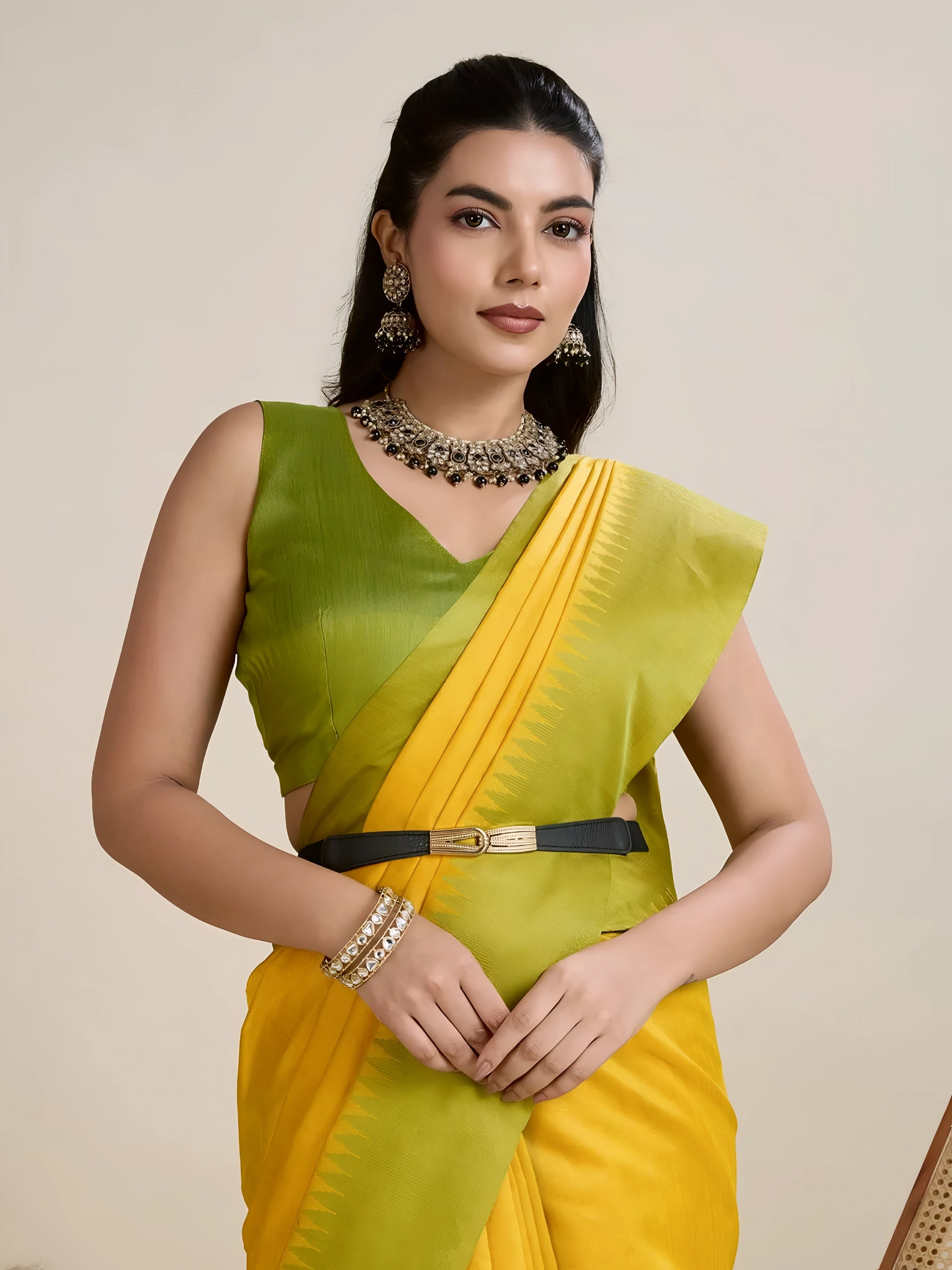 Woman wearing a yellow silk saree with a contrasting green border and pallu, paired with a matching blouse and traditional jewelry, elegantly posing beside a wooden chair in a minimalist studio setting with neutral decor, ceramic vases, and pampas grass.