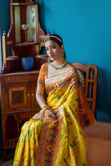 Woman wearing a vibrant yellow silk saree with multicolor geometric patterns and a matching blouse, accessorized with traditional jewelry, elegantly posing beside a wooden chair in a stylish room with teal blue walls, wooden bed, and decorative accents.