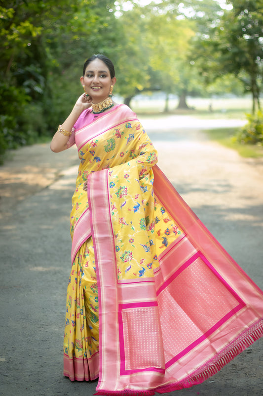 Woman wearing a yellow silk saree with pink borders and multicolor floral motifs, paired with a pink blouse and traditional jewelry, standing gracefully on a tree-lined garden path with soft natural light and lush greenery in the background.
