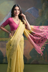 Woman wearing a light yellow saree with a contrasting pink pallu featuring floral embroidery, paired with a matching pink blouse and traditional jewelry, standing in front of a painted mountain and forest backdrop.