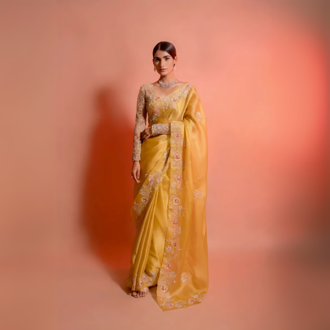 Woman wearing a yellow silk saree with floral embroidery and a matching embellished blouse, posing in a studio with a peach gradient background.