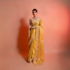 Woman wearing a yellow silk saree with floral embroidery and a matching embellished blouse, posing in a studio with a peach gradient background.