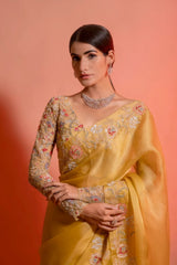 Woman wearing a yellow silk saree with floral embroidery and a matching embellished blouse, posing in a studio with a peach gradient background.