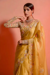 Woman wearing a yellow silk saree with floral embroidery and a matching embellished blouse, posing in a studio with a peach gradient background