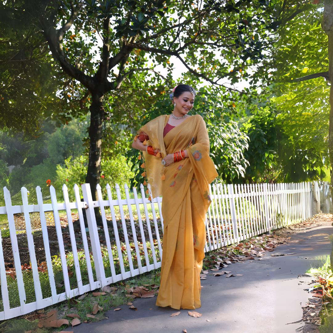 Woman wearing a mustard yellow silk saree with subtle floral embroidery and a contrasting maroon blouse, accessorized with traditional jewelry and bangles, standing beside a white picket fence in a sunlit garden with lush greenery and trees in the background.