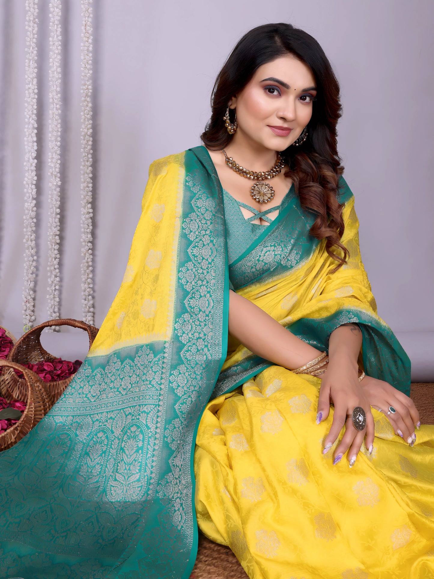  Woman wearing a vibrant yellow silk saree with an ornate turquoise blue border and matching blouse, featuring subtle floral motifs and traditional zari work. She poses gracefully in a contemporary studio adorned with woven baskets filled with rose petals, against a neutral backdrop with decorative vertical garlands.