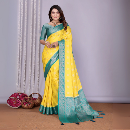  Woman wearing a vibrant yellow silk saree with an ornate turquoise blue border and matching blouse, featuring subtle floral motifs and traditional zari work. She poses gracefully in a contemporary studio adorned with woven baskets filled with rose petals, against a neutral backdrop with decorative vertical garlands.